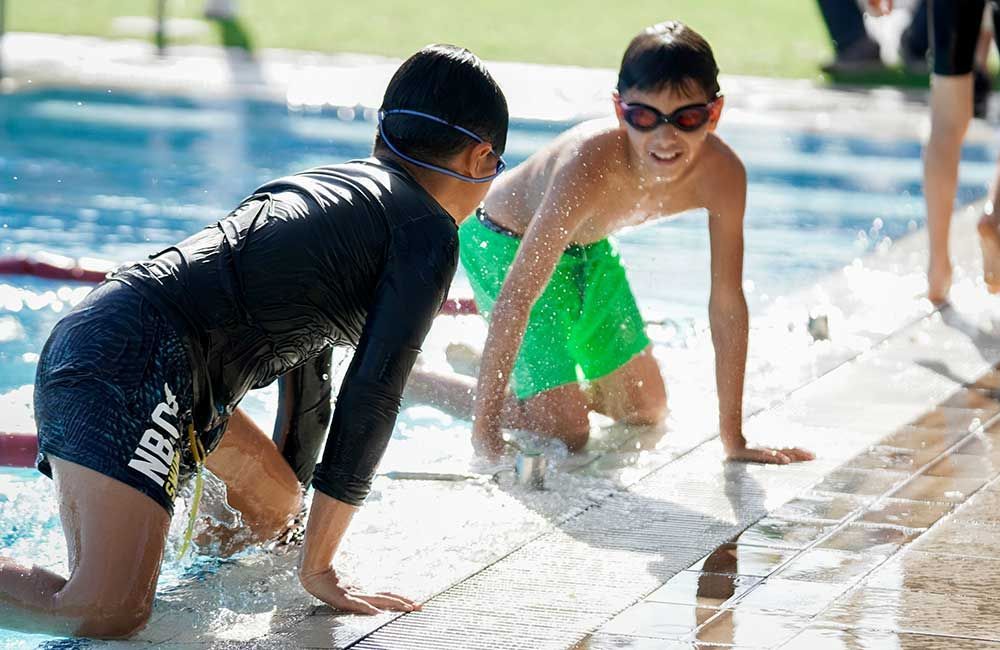 Two young boys are kneeling on the edge of a swimming pool.