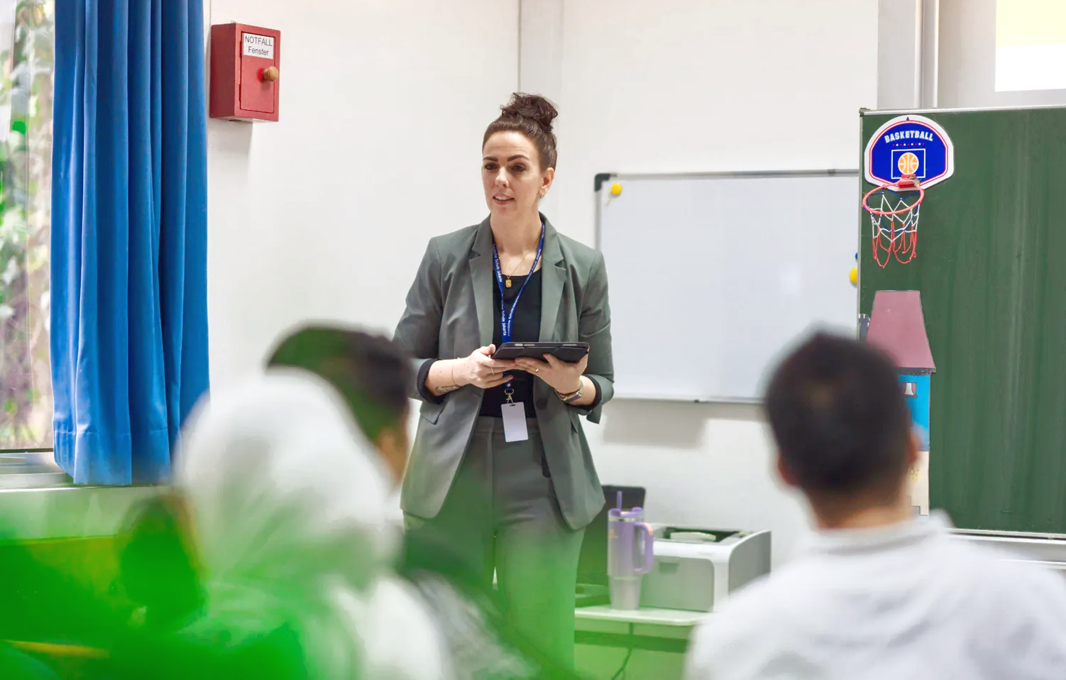 Woman in gray suit presenting in classroom, holding tablet. Students are blurred in the foreground.