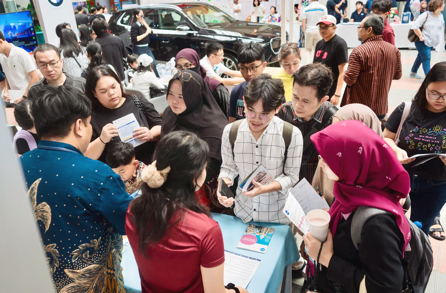 People gather at a booth, likely an event. Some look at brochures. A BMW car is visible in the background.
