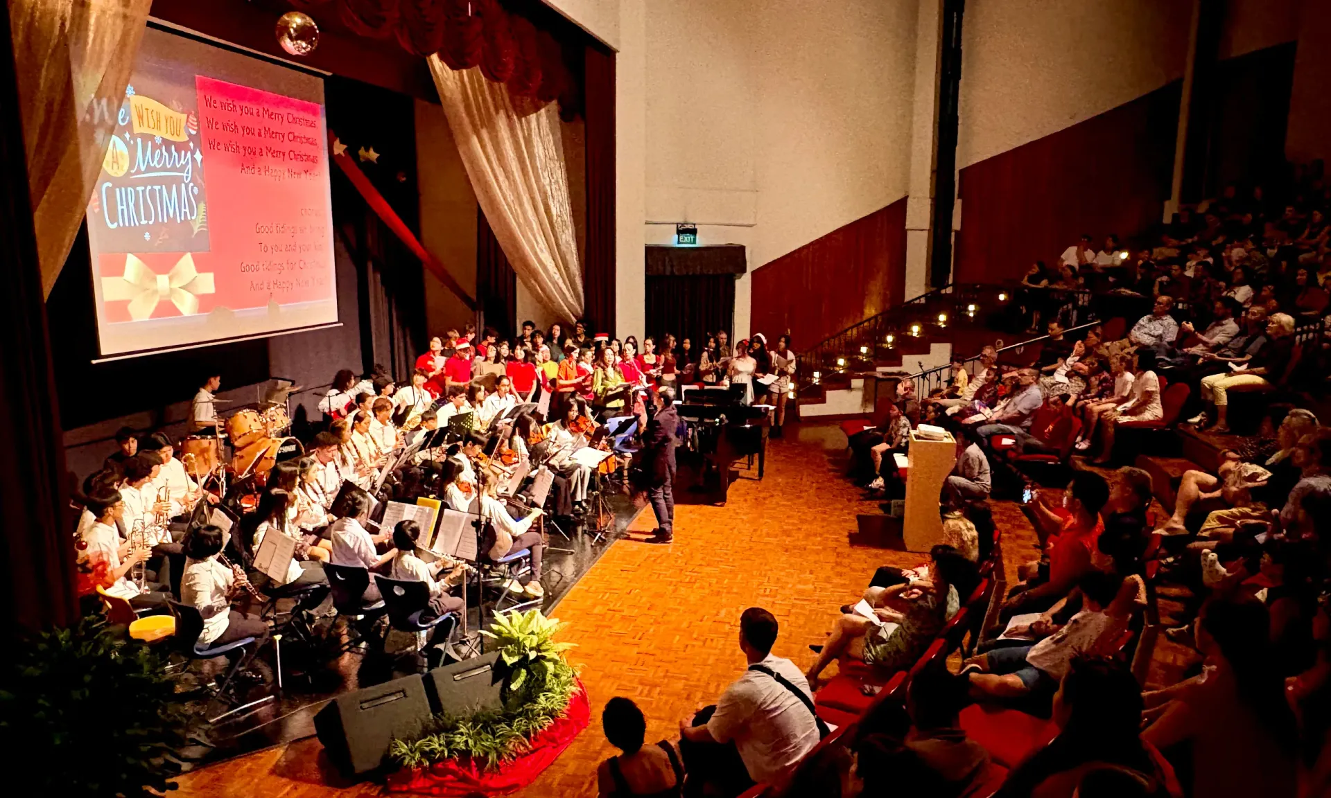 Audience in an auditorium watches performers on stage with a screen displaying text. Stage is decorated.