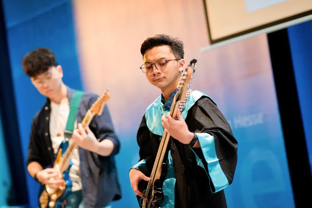 Two men in graduation gowns are playing guitars on a stage.
