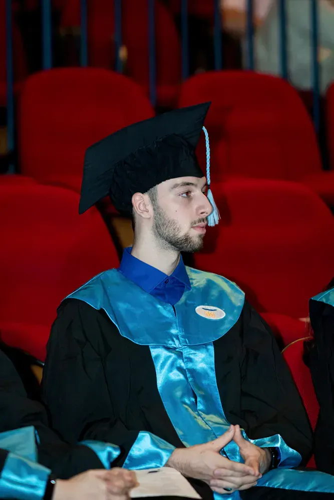 A man in a graduation cap and gown is sitting in a row of red chairs.