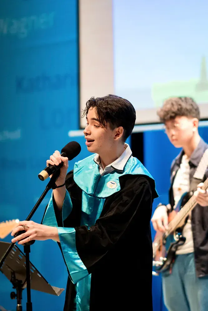 A man in a graduation gown is singing into a microphone while another man plays a guitar.