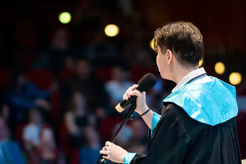 A man in a graduation gown is giving a speech into a microphone.
