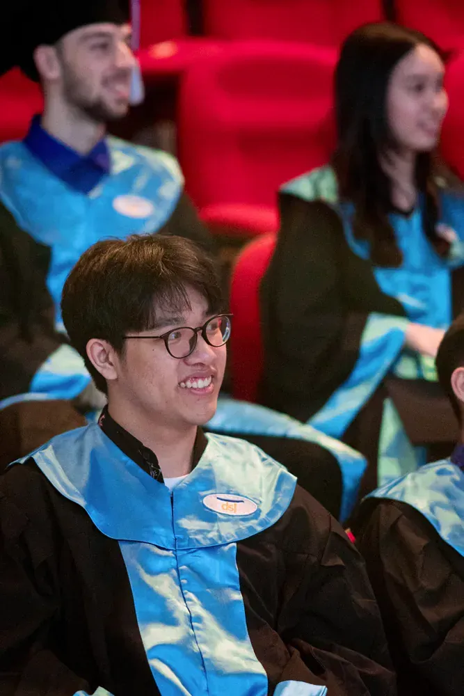 A group of graduates are sitting in a auditorium and smiling.