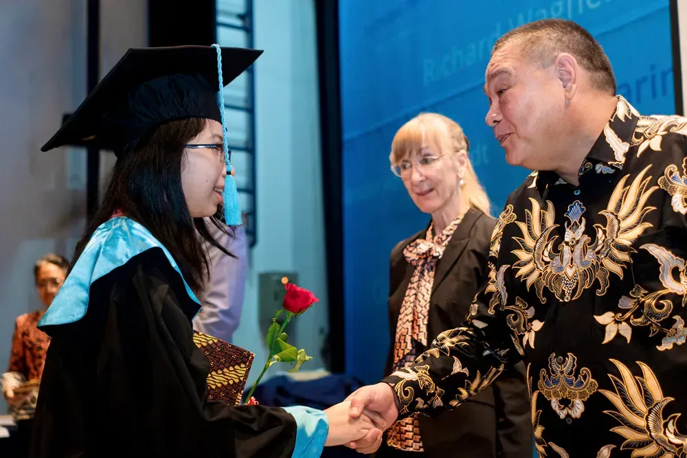 A woman in a graduation cap and gown is shaking hands with a man in a floral shirt.