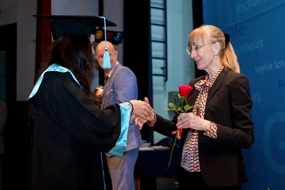 A woman in a graduation cap and gown is shaking hands with another woman holding a red rose.