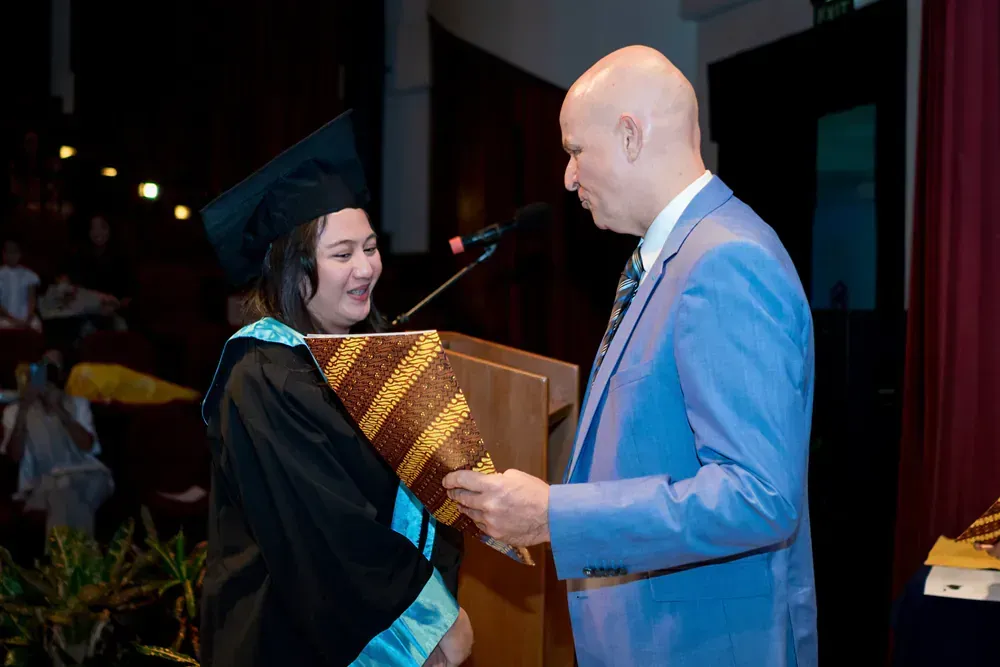 A man in a suit is giving a diploma to a woman in a graduation cap and gown.