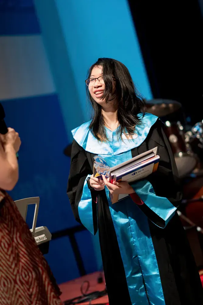 A woman in a graduation gown is holding a book and smiling