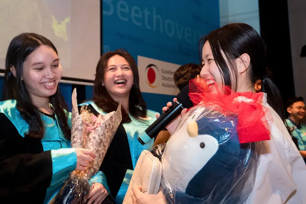A woman is holding a stuffed penguin and a bouquet of flowers while talking into a microphone.