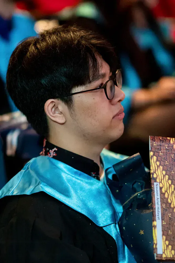 A young man wearing glasses and a graduation gown is sitting in a classroom.