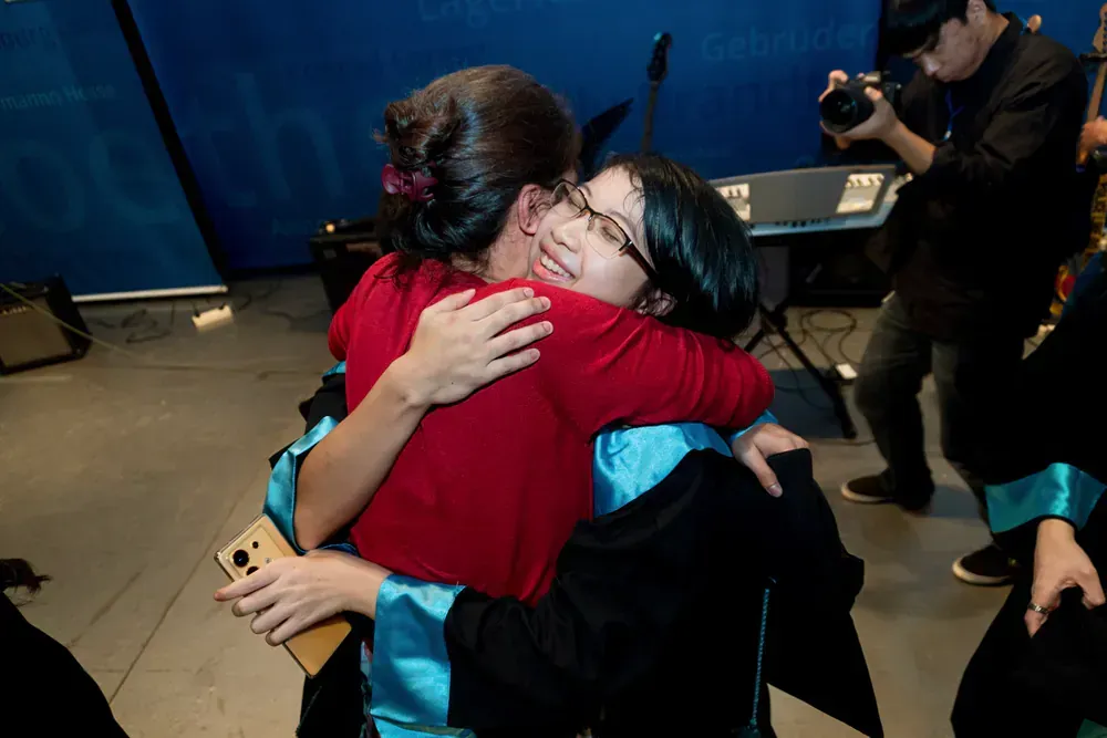 A woman in a graduation gown is hugging another woman in a red shirt.