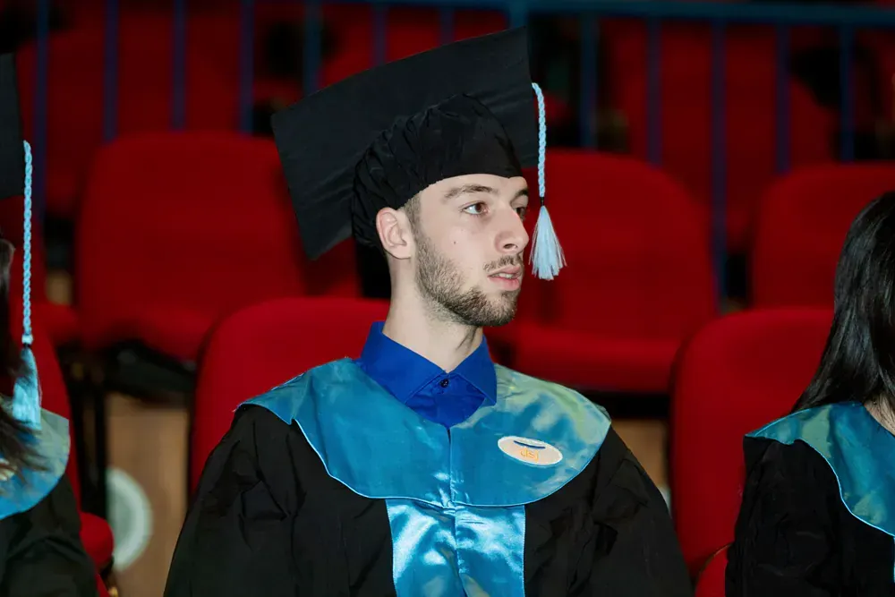 A man in a graduation cap and gown is sitting in a row of red chairs.