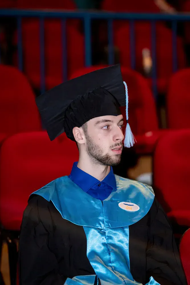 A man in a graduation cap and gown is sitting in a row of red chairs.