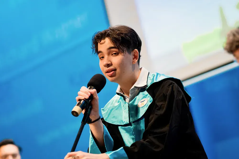 A young man in a graduation gown is speaking into a microphone.
