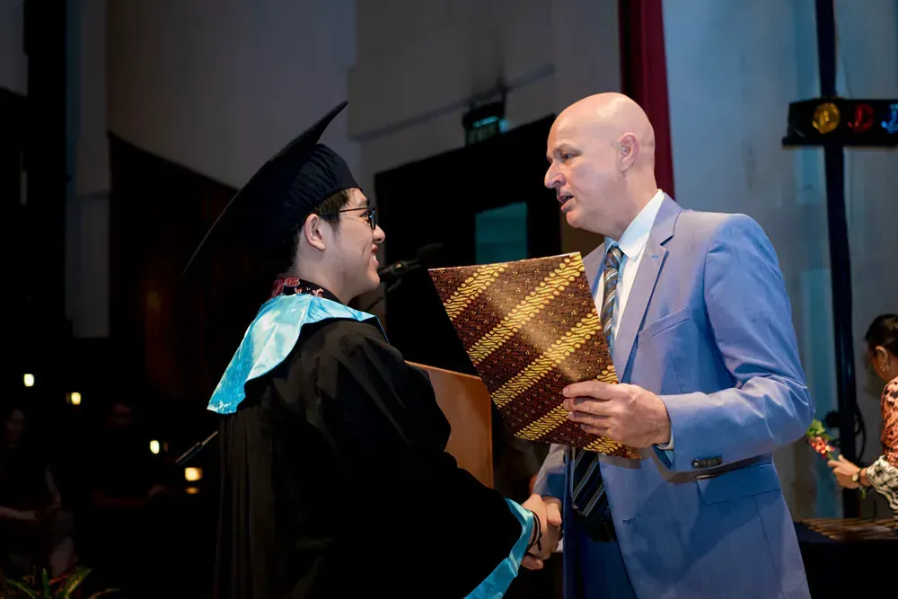 A man in a suit is shaking hands with a graduate in a cap and gown.