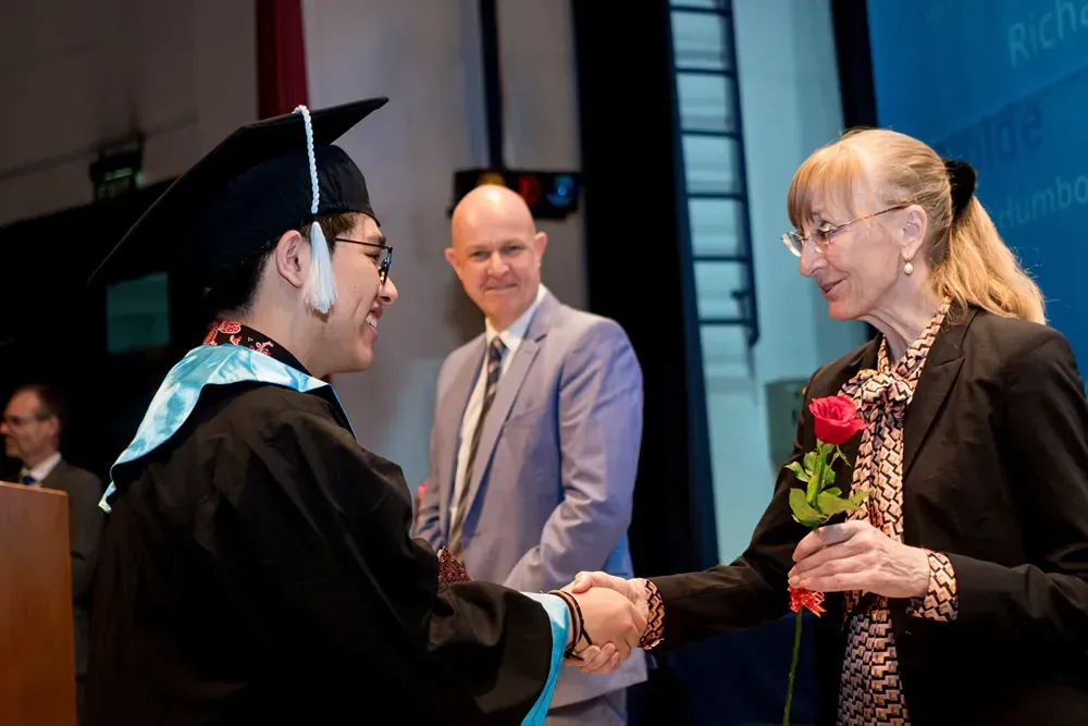 A man in a graduation cap and gown is shaking hands with a woman holding a rose.