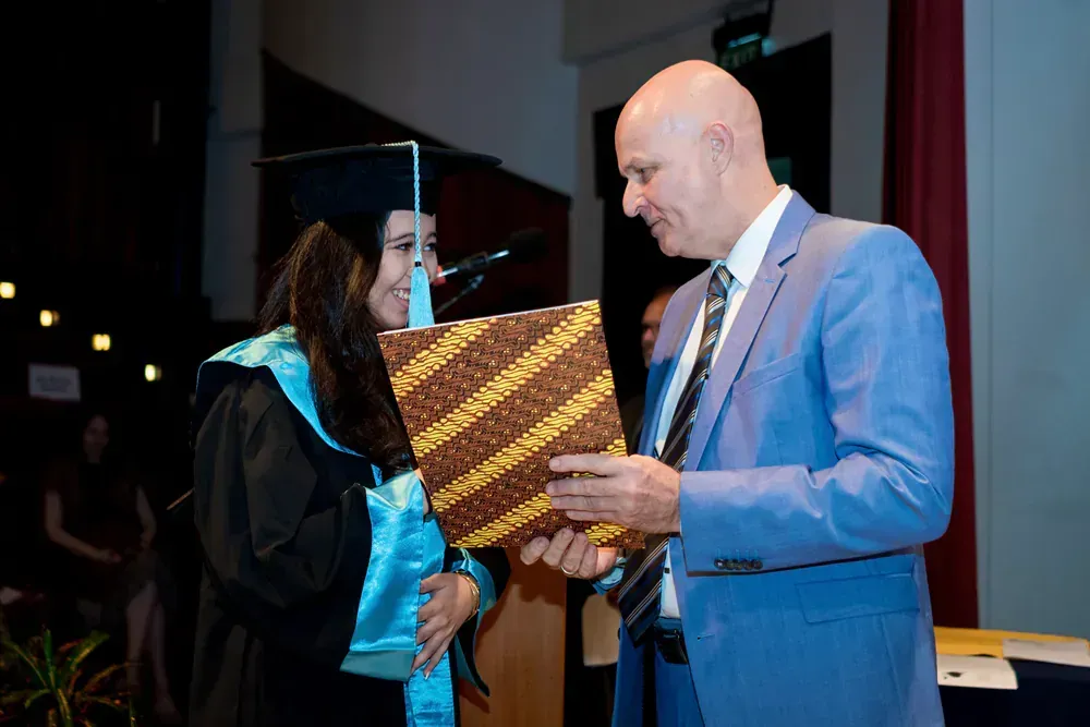 A man in a suit is giving a diploma to a woman in a graduation cap and gown.