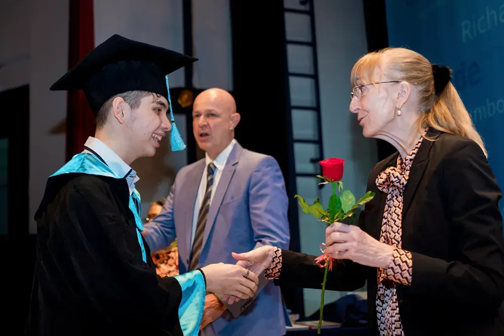 A man in a graduation cap and gown is shaking hands with a woman holding a rose.