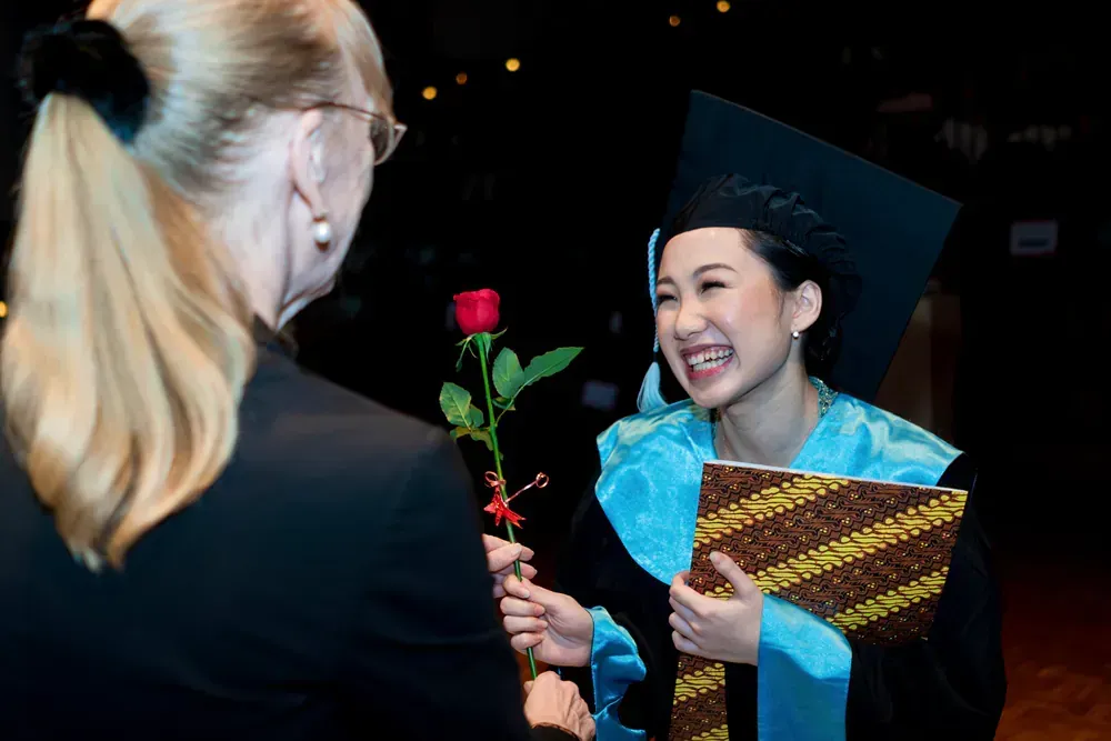 A woman is giving a rose to a woman in a graduation cap and gown.