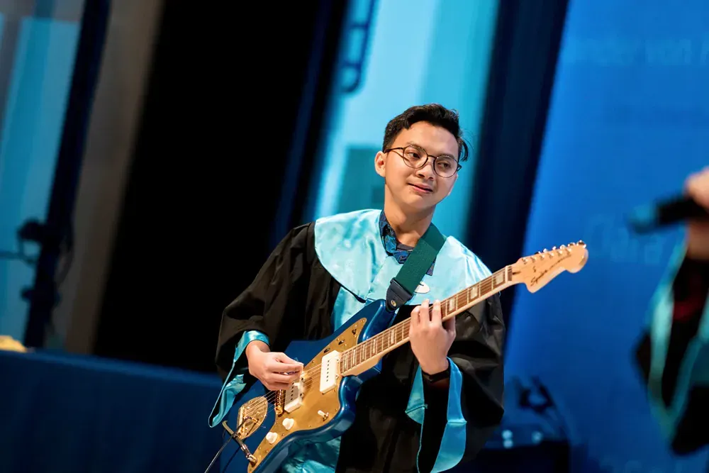 A man in a graduation cap and gown is playing a guitar on a stage.