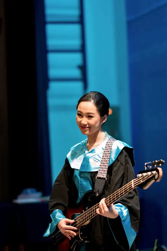 A woman in a graduation gown is playing a guitar on stage.