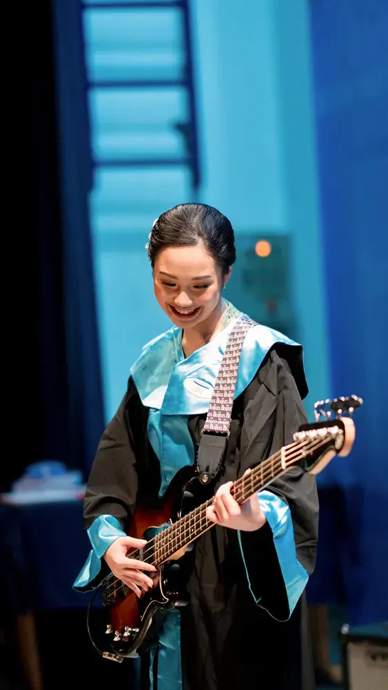 A woman in a graduation gown is playing a guitar on a stage.