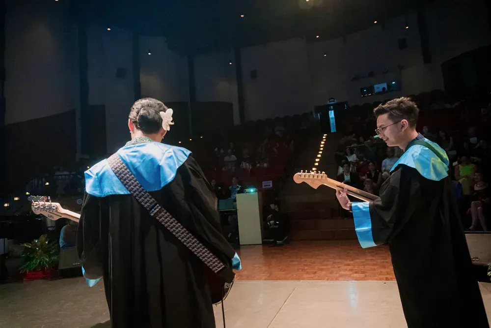 Two people in graduation gowns are playing guitars on a stage.