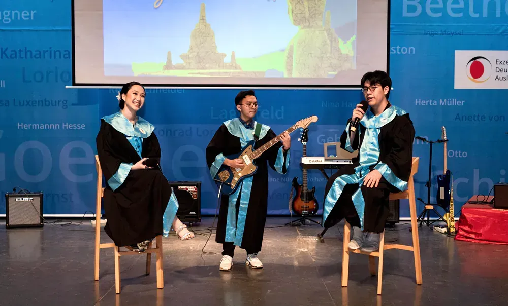 Three people in graduation gowns are sitting in chairs on a stage playing guitars.