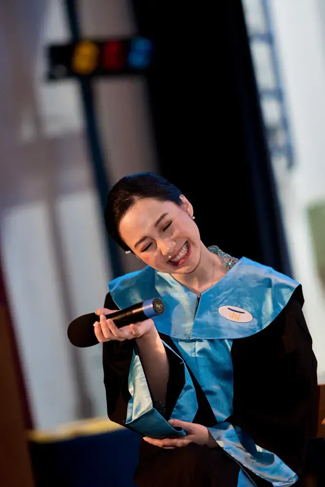 A woman in a graduation gown is holding a microphone and smiling.