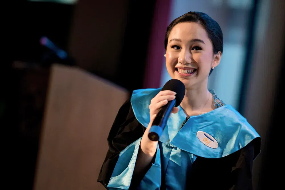 A woman in a graduation gown is holding a microphone and smiling.
