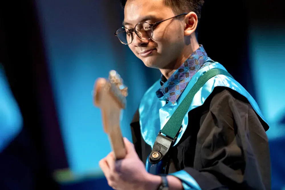 A man in a graduation gown is playing an electric guitar.