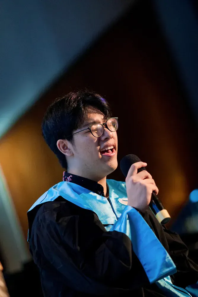 A young man in a graduation gown is singing into a microphone.