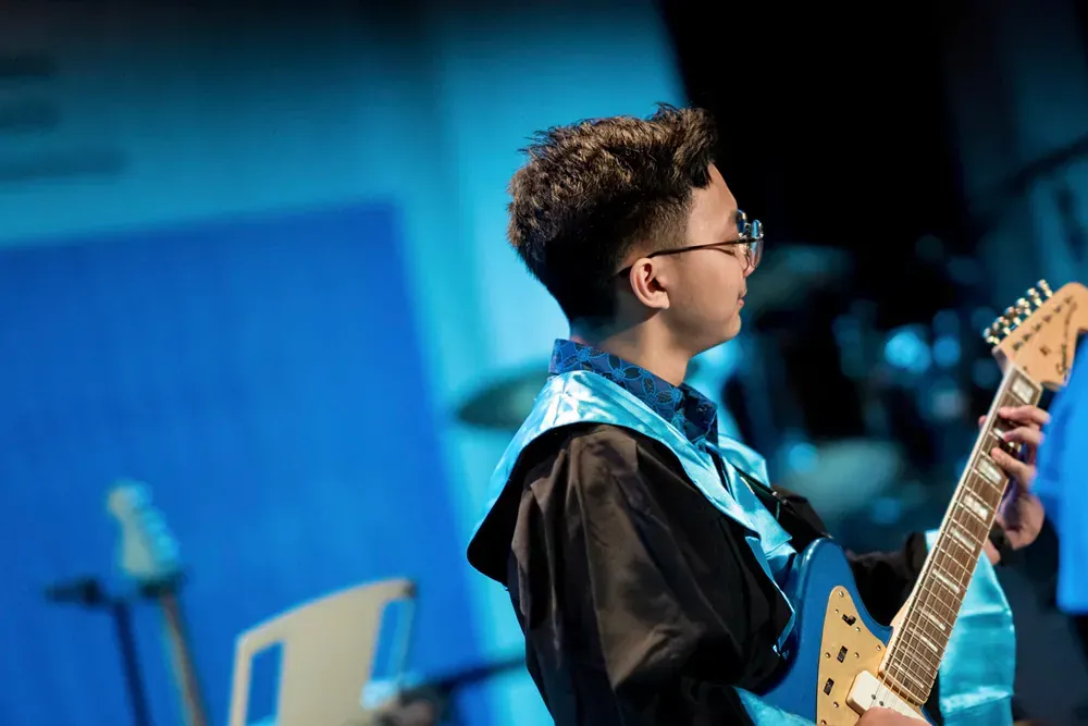 A young man in a graduation cap and gown is playing an electric guitar.