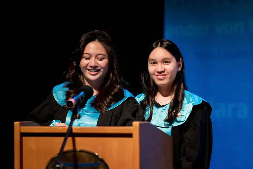 Two women in graduation gowns are standing at a podium with microphones.