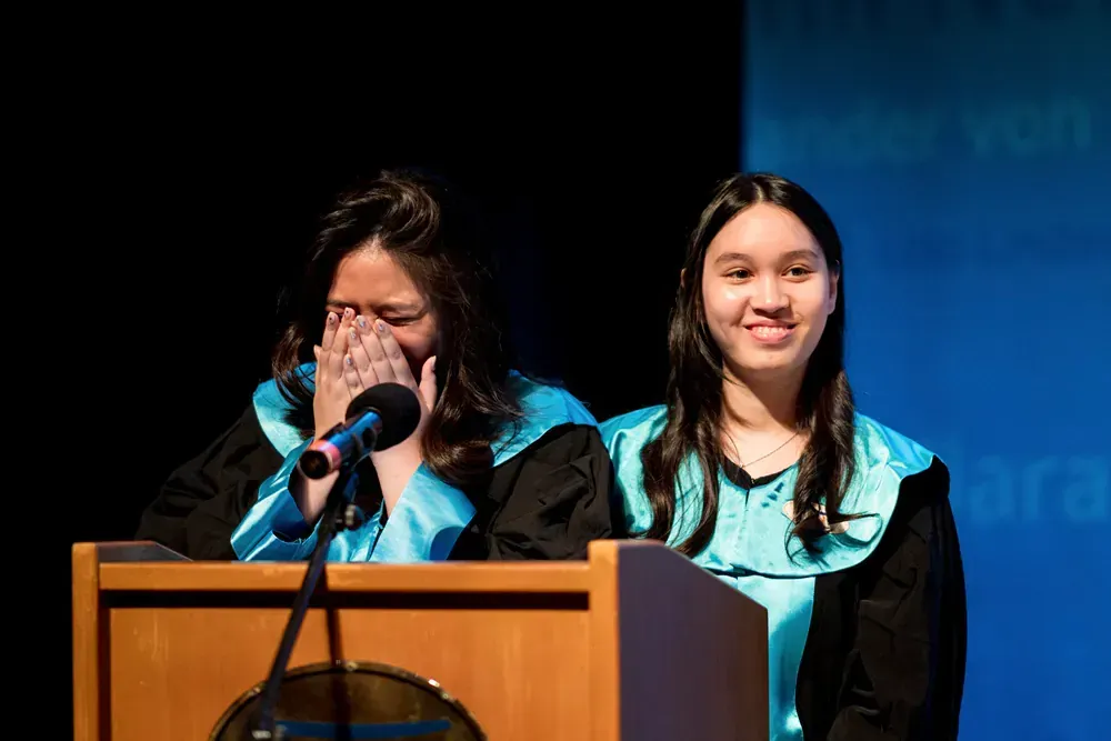 Two women in graduation gowns are standing at a podium covering their faces with their hands.
