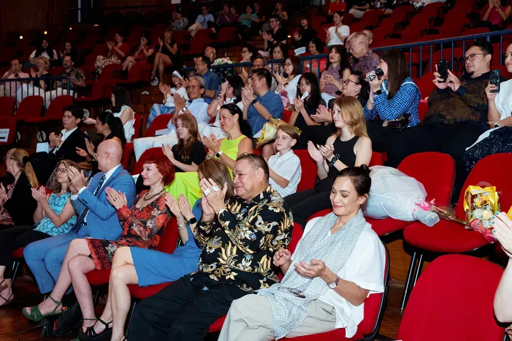 A large group of people are sitting in red chairs in a theater applauding.