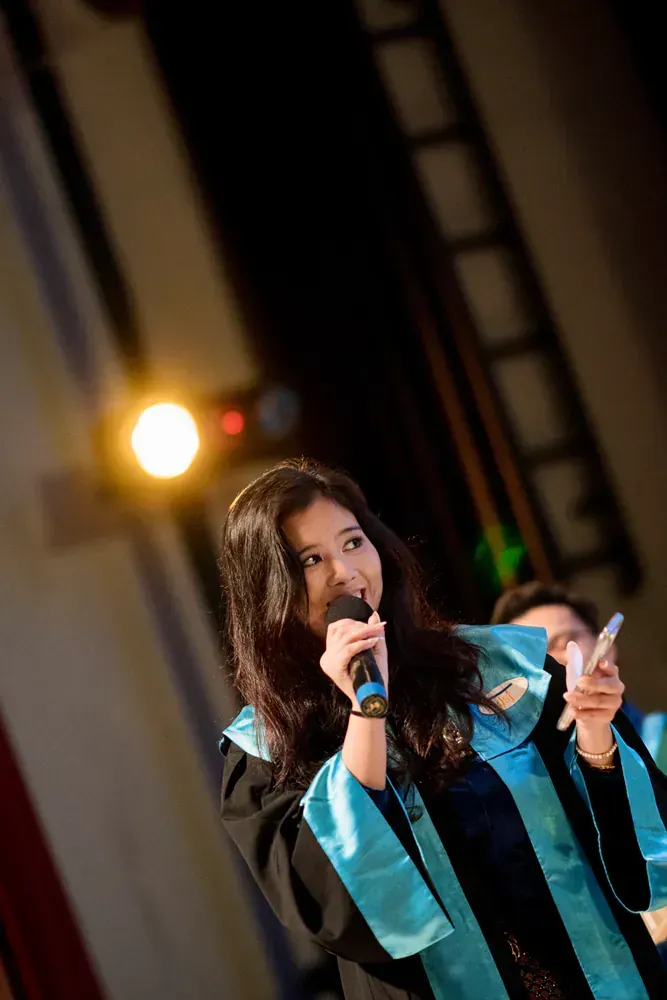 A woman in a graduation gown is holding a microphone and a cell phone.