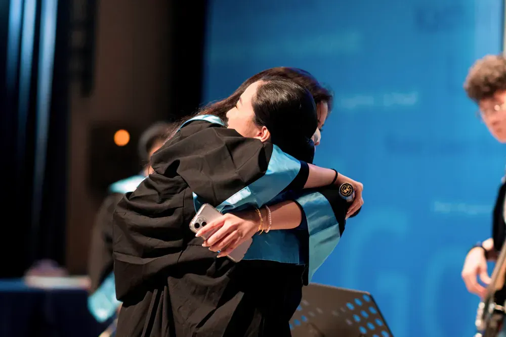 Two women in graduation gowns are hugging each other on a stage.