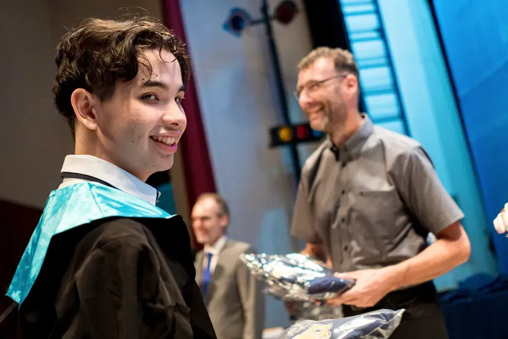 A young man in a graduation cap and gown is standing next to a man holding a bag of clothes.