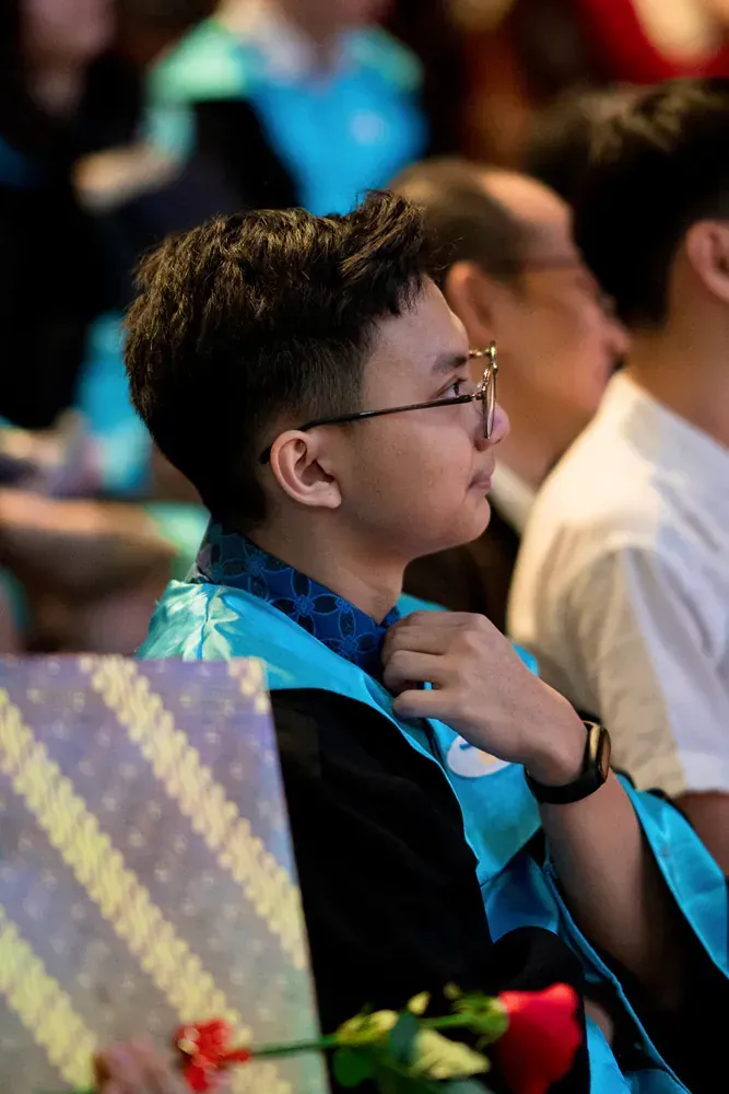 A young man in a graduation cap and gown is sitting in a crowd of people.