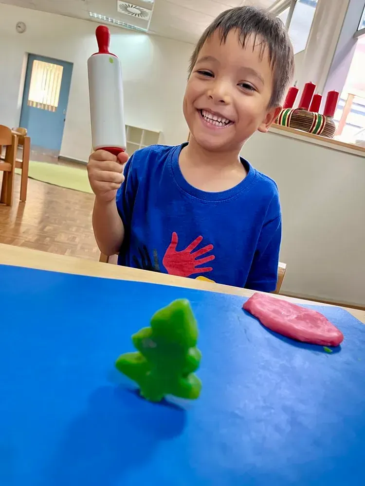 A young boy is sitting at a table holding a rolling pin and smiling.