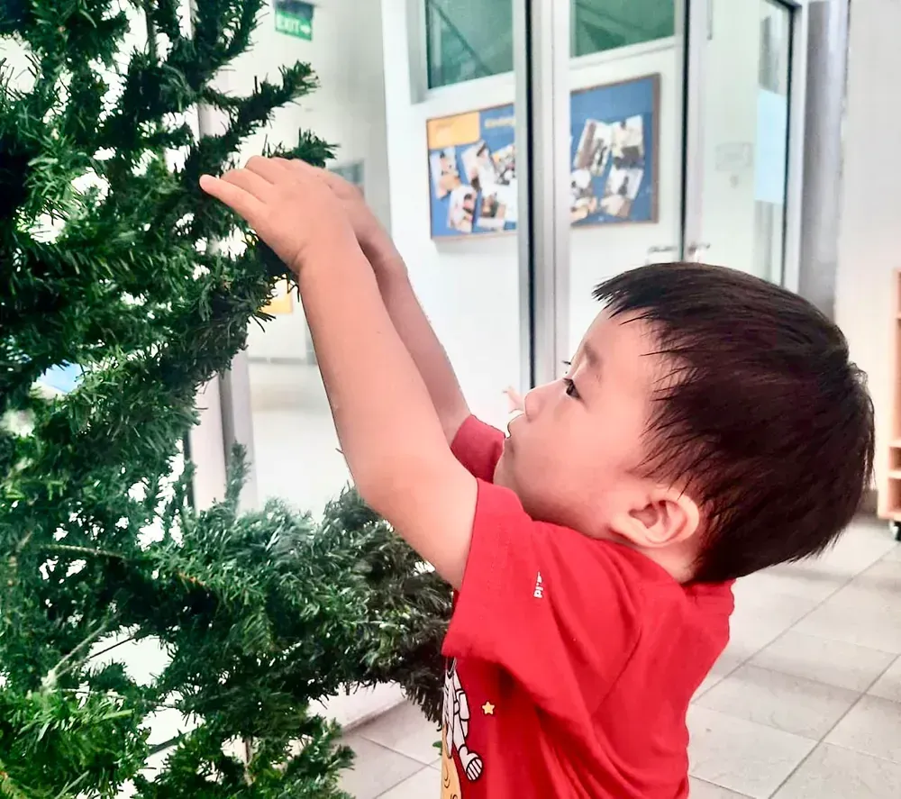 A little boy in a red shirt is decorating a christmas tree