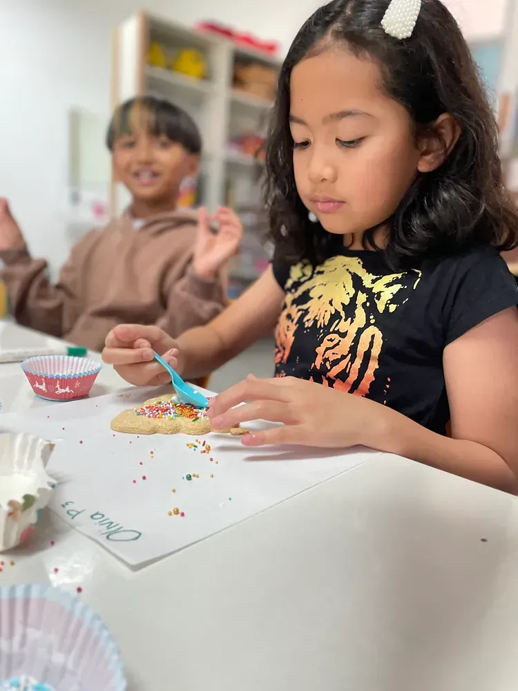 A little girl is sitting at a table decorating a cookie with sprinkles.