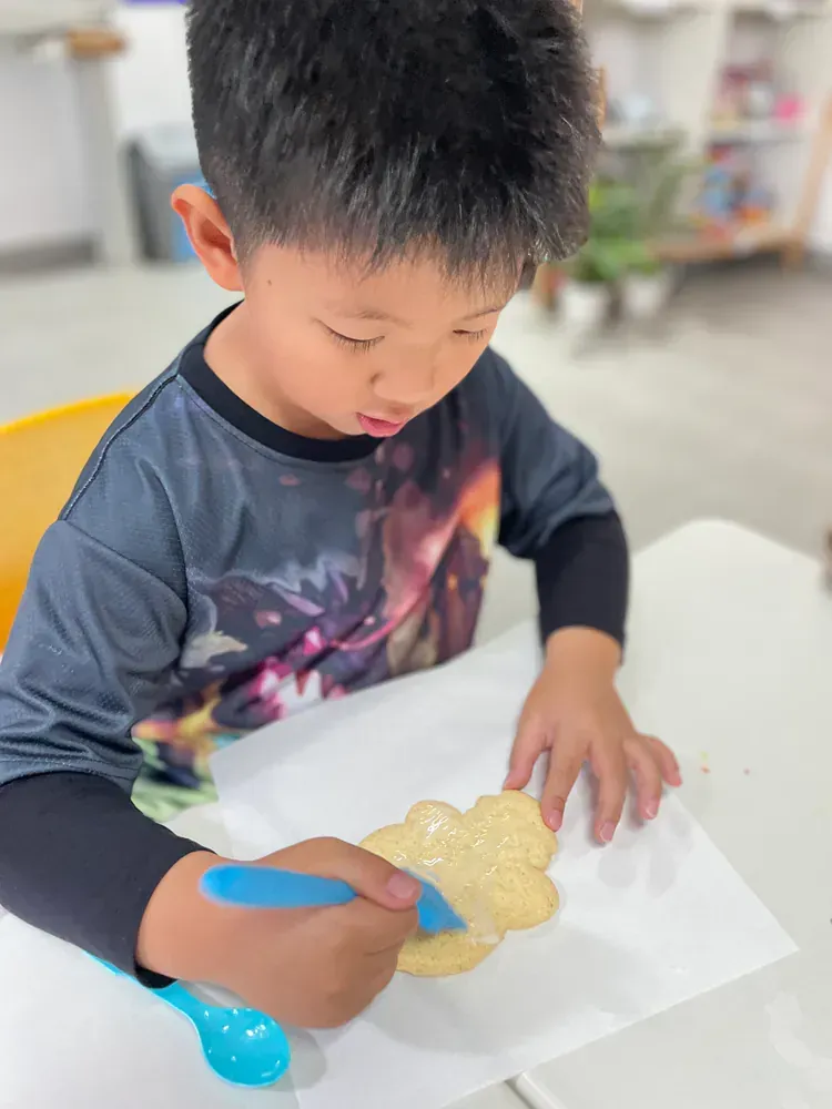 A young boy is sitting at a table holding a cookie and a spoon.