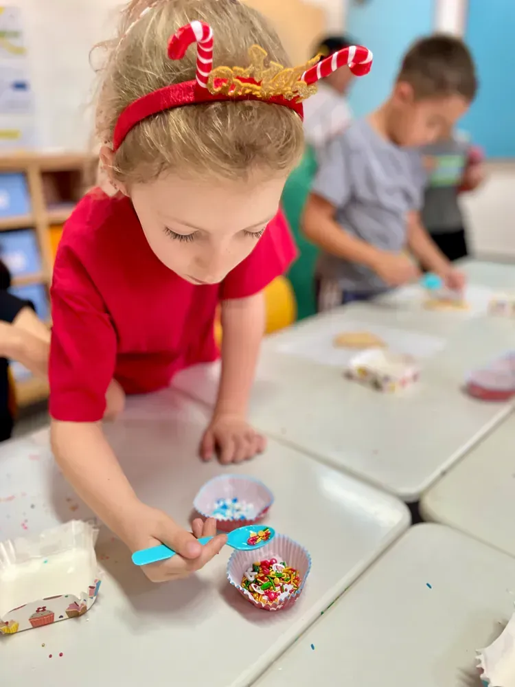 A little girl wearing a candy cane headband is playing with sprinkles at a table.