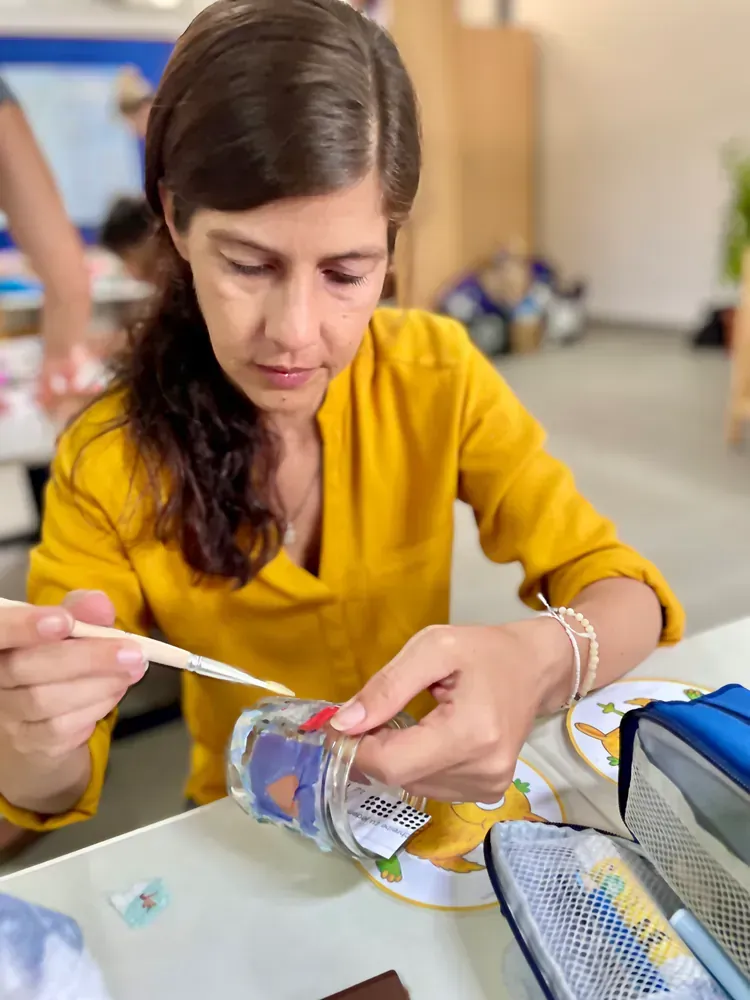 A woman in a yellow shirt is sitting at a table painting a bottle.