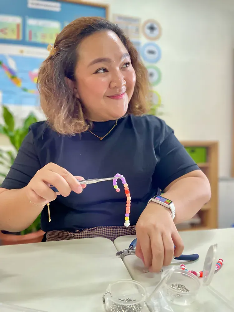 A woman is sitting at a table making a bracelet with beads.