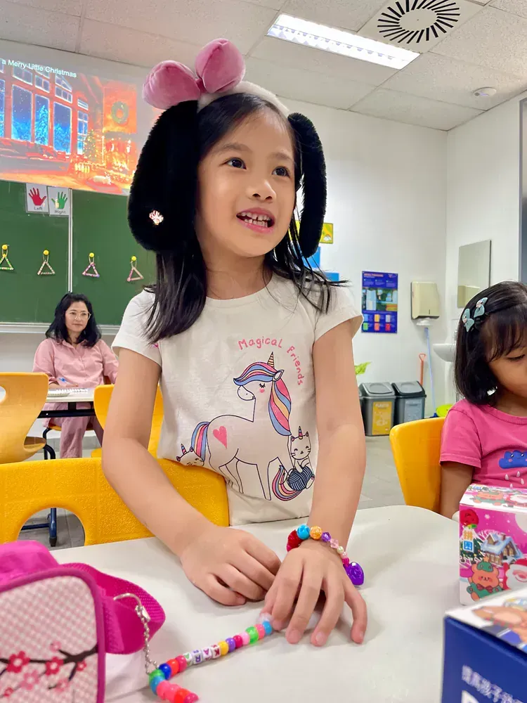 A little girl wearing a unicorn shirt is sitting at a table in a classroom.