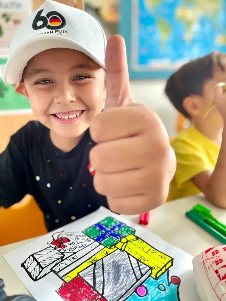 A young boy is giving a thumbs up while sitting at a table with a drawing on it.